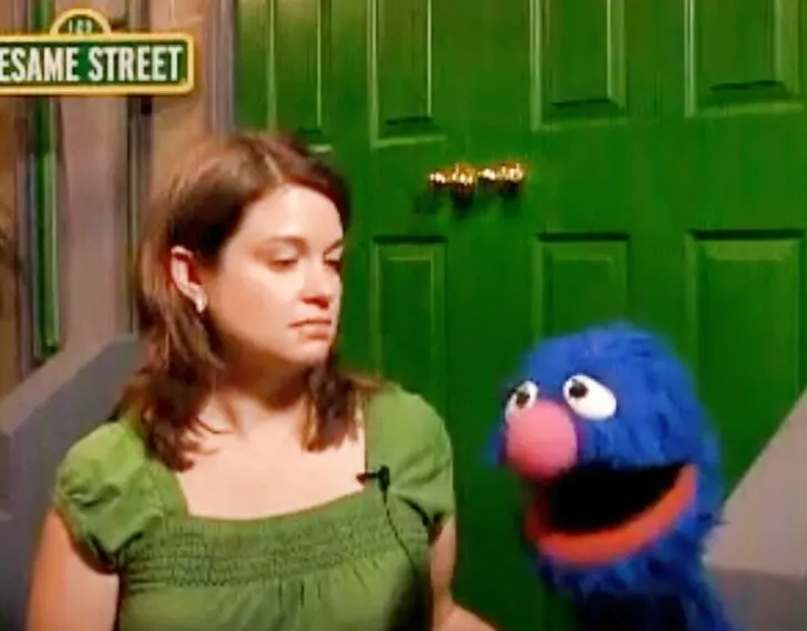 Amy Oztan and Grover the Muppet, sitting on a stoop on a Sesame Street set.