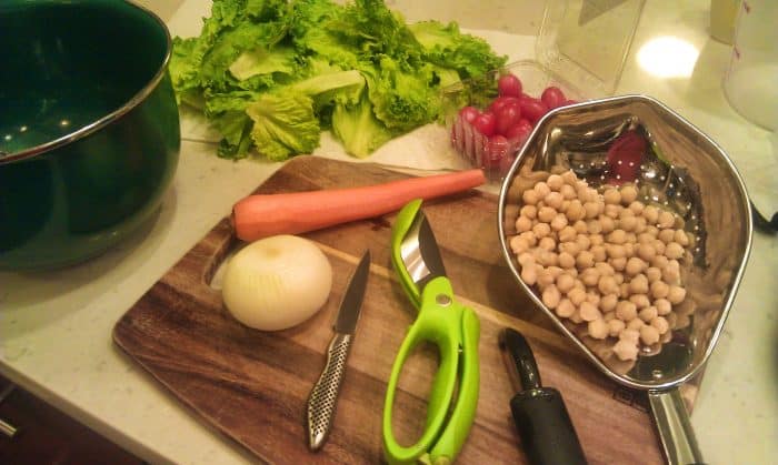 A wooden cutting board with lettuce, carrot, onion, chickpeas, a paring knife, a Toss and Chop, and a peeler.