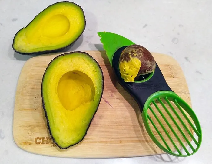 An avocado slicer on a small cutting board with two halves of an avocado.