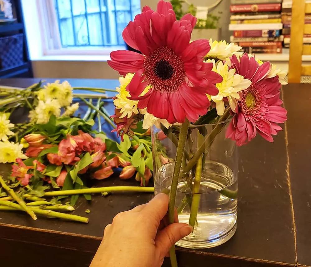 A hand holding some flowers in front of a vase.