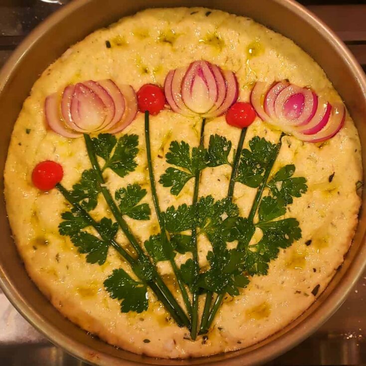 Focaccia dough in a cake pan decorated with vegetables and herbs.